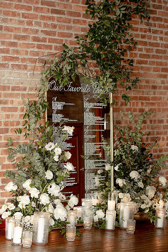Wedding seating chart mirror sign with calligraphy lettering on a gold-framed mirror, accented by greenery garland and candles against a brick wall