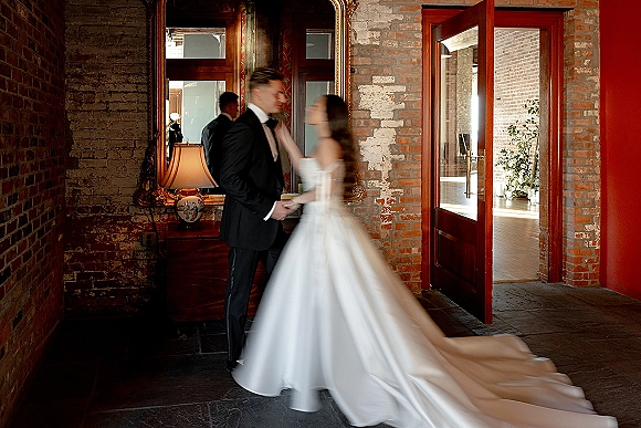 Wedding couple portrait of bride and groom holding hands in a brick hallway by an ornate framed mirror, her long train trailing behind