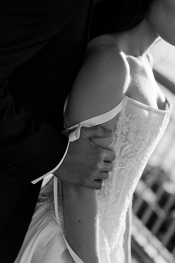 Wedding couple portrait in a close embrace, groom hugging bride at the waist, highlighting lace bodice and satin ribbon against a soft blur background