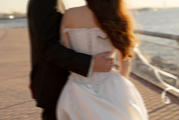 Couple portrait of bride and groom in embrace, her strapless gown and veil trailing, on a beach boardwalk by the ocean at sunset