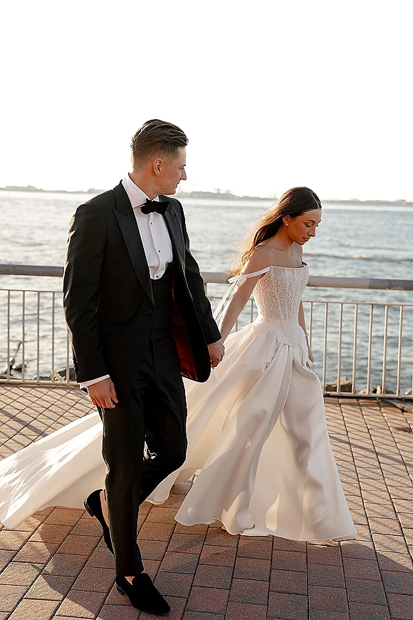 Couple portrait of bride and groom walking hand in hand, her long train flowing by an ocean waterfront railing at sunset