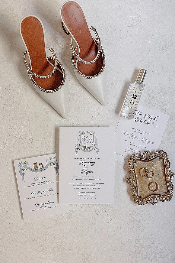 Invitation suite wedding invitation flat lay with monogram crest cards, gold rings on ornate tray, and white rhinestone heels on textured surface
