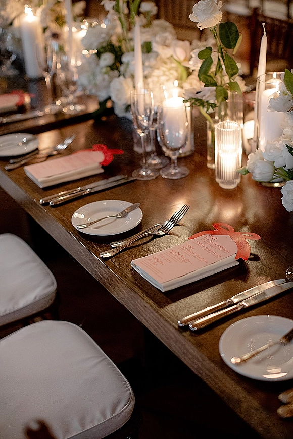 Reception tablescape with wedding table centerpiece of white florals, taper and votive candles, and red ribbon menus on a wood table in dim light