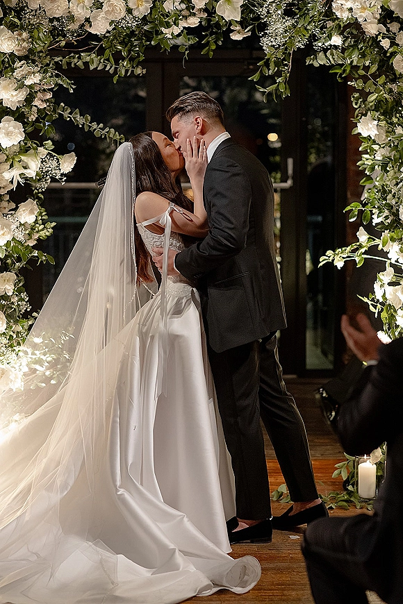 Wedding kiss as bride and groom embrace under a white floral arch, veil trailing over wooden floor, candle accents by glass doors