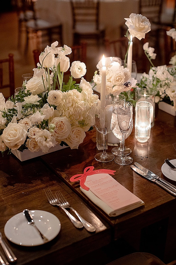 Reception tablescape with a wedding table centerpiece of white flowers, taper candles, ribbed glass votives, and glassware on a wooden table in a dim room