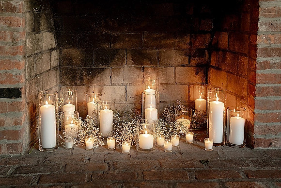 Wedding candle decor with pillar candles in hurricanes and votives nestled in greenery, clustered on a brick fireplace hearth for warm glow