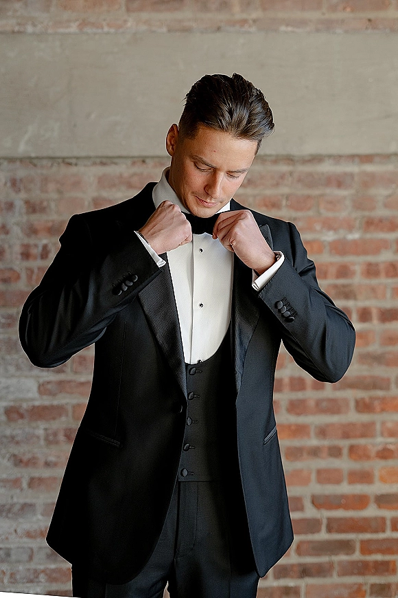 Groom portrait in a classic black tuxedo with bow tie and waistcoat, posing against a textured brick wall backdrop