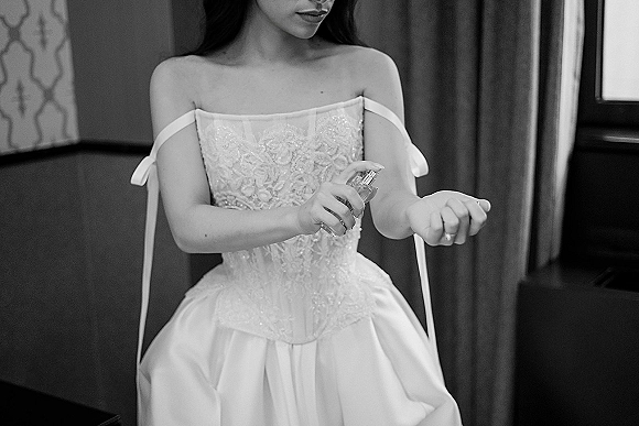 Bridal getting ready as the bride sprays perfume in an off the shoulder corset lace gown by a window with curtains and chair