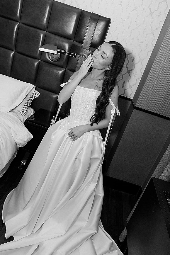 Bridal portrait in black and white of a bride seated on a hotel bed, looking away, wearing an off-shoulder gown with ribbon straps