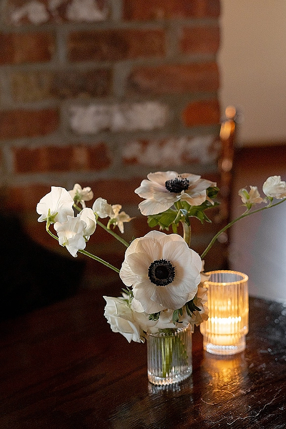 Wedding centerpiece with white anemone flowers in a bud vase beside a ribbed glass votive candle on a wooden table against brick wall