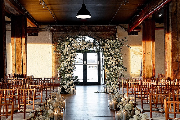 Ceremony aisle decor with a candle lined aisle leading to a white floral arch, greenery and Chiavari chairs in an industrial brick venue