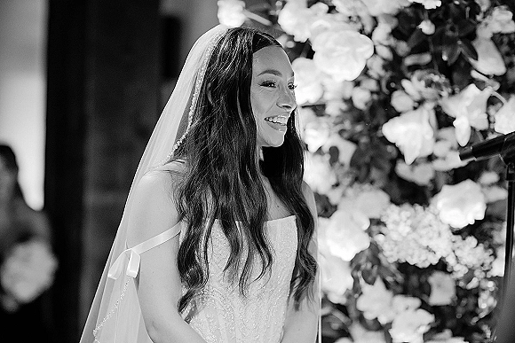 Bride portrait of a laughing bride at a microphone in a beaded strapless gown, with wedding veil and floral backdrop, guests blurred behind