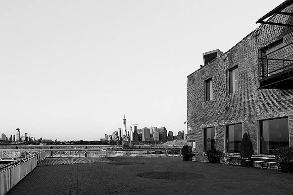 Wedding venue exterior of an industrial wedding venue with potted shrubs and benches along a riverfront promenade and city skyline backdrop