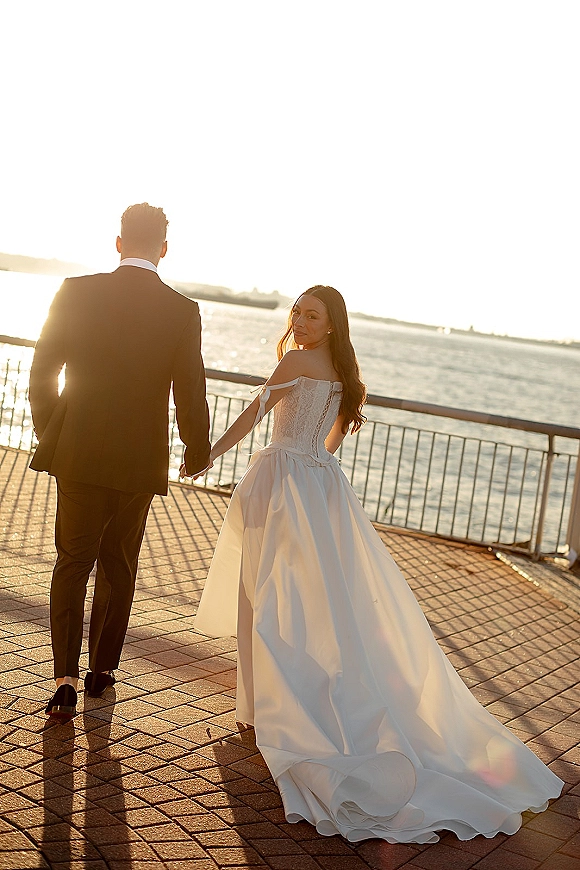 Couple portrait of bride and groom walking away, holding hands on a brick waterfront promenade with sun flare and ocean horizon behind