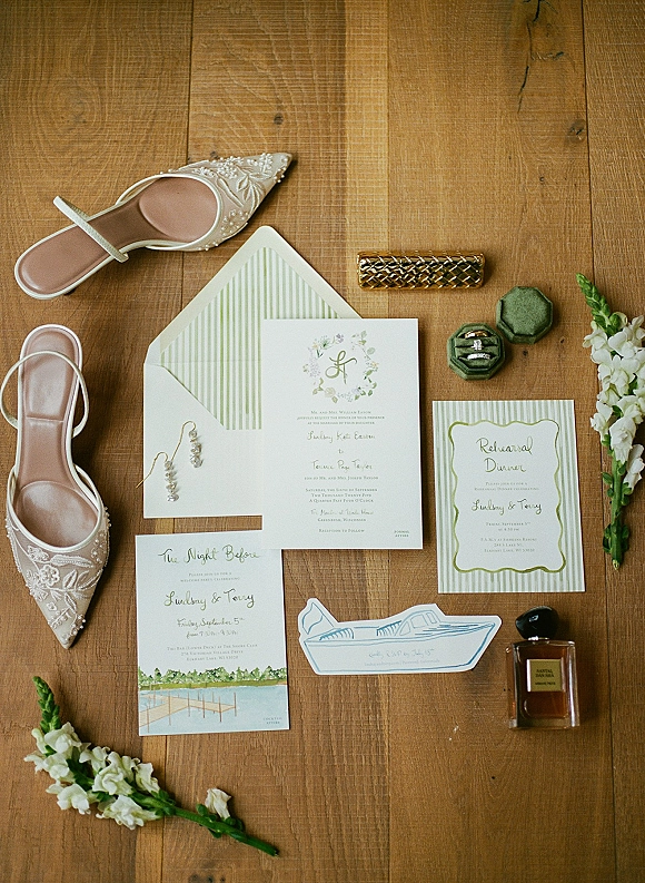 Wedding invitation suite with green wedding invitations, striped envelope liner, wax seal, rings, earrings, perfume and shoes on a wood table