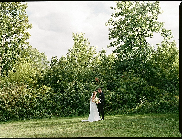 Couple portrait of bride and groom embracing, bride holding a white bouquet and veil flowing, on a green lawn with trees and clouds