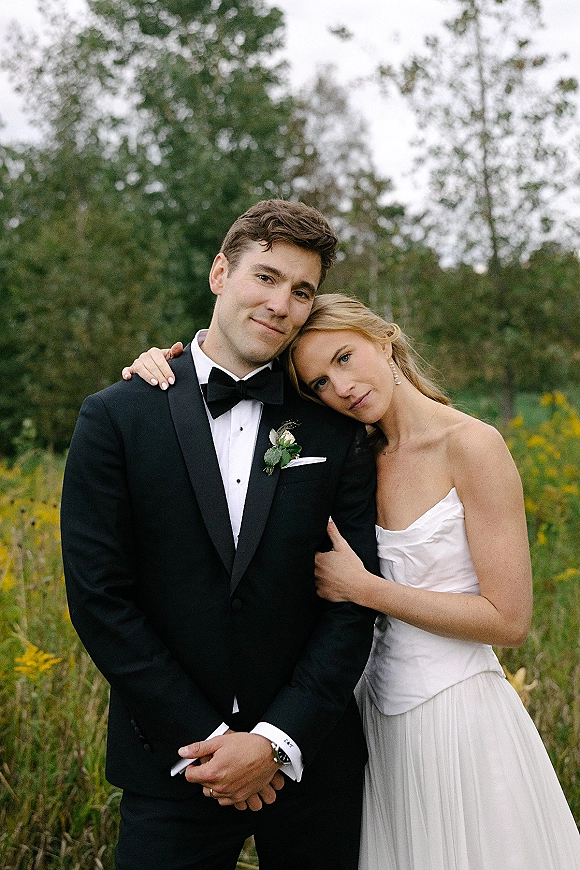 Couple portrait of bride resting head on groom in a wildflower meadow, wearing a strapless wedding dress and black tuxedo under overcast sky