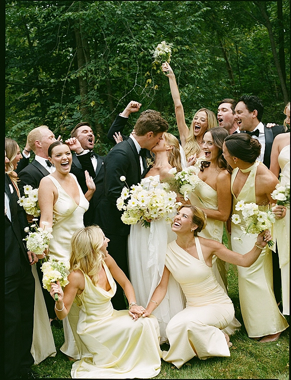 Wedding party photo of bride and groom kissing as bridesmaids in yellow dresses cheer with white bouquets on a garden lawn