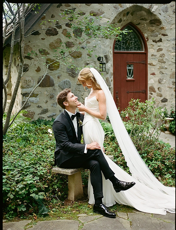 Couple portrait of bride touching groom’s face as he sits on a stone bench by a church wall, her long veil trailing behind