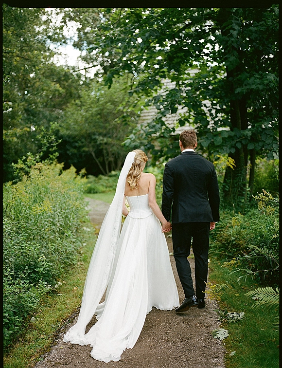 Couple walking away hand in hand, newlyweds walking hand in hand along a tree-lined garden path, bride’s long veil trailing in sunlight