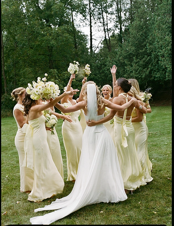 Bridal party moment as the bride with bridesmaids in butter yellow dresses forms a circle, raising white bouquets on a garden lawn