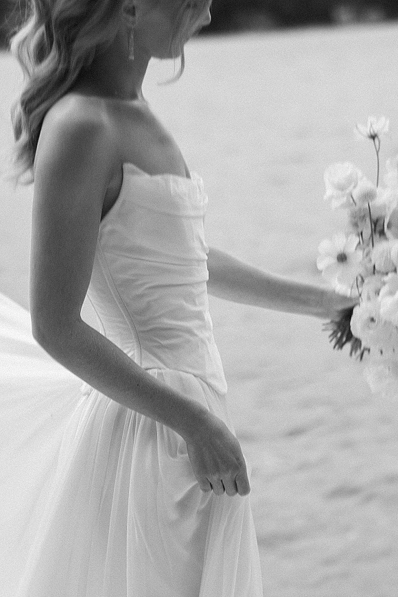 Bridal portrait in black and white of a bride in a strapless wedding dress holding a bouquet by the shoreline with water and sky behind