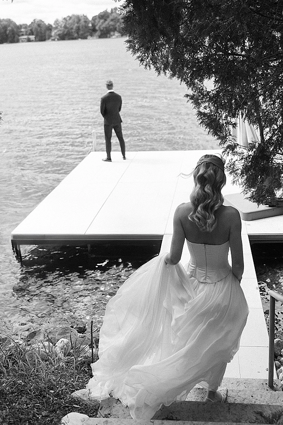 Wedding first look as bride approaches groom waiting on a dock, holding her tulle train by the railing with lake and trees behind