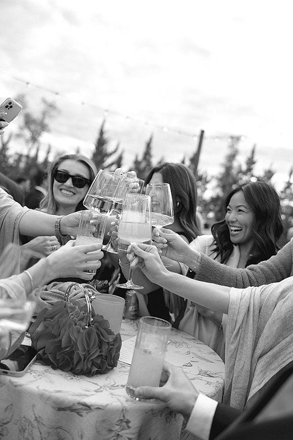 Wedding toast as friends clink wine and mixed drink glasses over a cocktail table with floral centerpiece, string lights and trees behind
