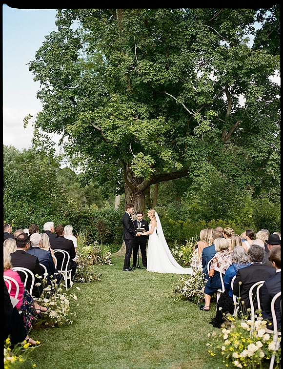 Wedding ceremony with bride and groom exchanging vows under a leafy tree arch, floral-lined aisle and white chairs in a garden setting