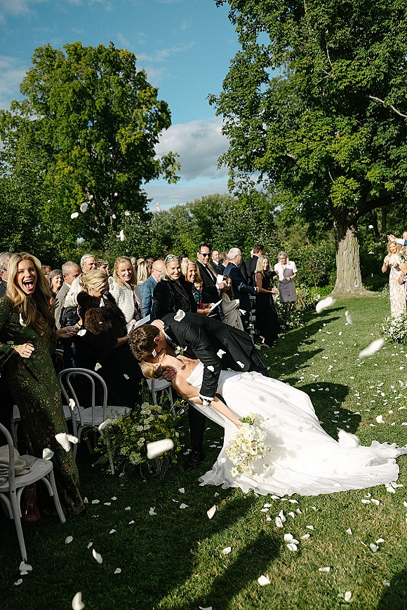 Wedding kiss moment as newlyweds dip in the aisle while guests cheer and white petals fly, long-train dress and bouquet in sunny garden