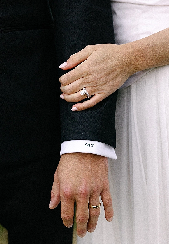 Wedding rings shown in an engagement ring close up as bride’s manicured hand rests on groom’s arm against a neutral backdrop