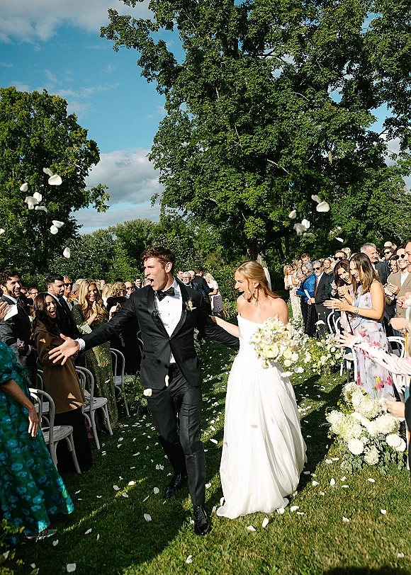 Wedding recessional as bride and groom walk the aisle hand in hand, bride holding bouquet amid flower petals and cheering guests in a garden setting