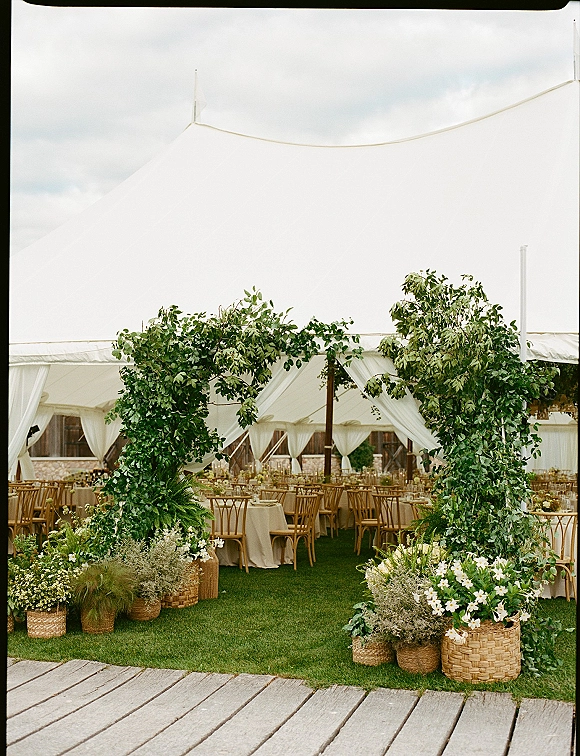 Wedding reception decor with a greenery entrance arch, woven baskets of white flowers and potted ferns under a draped sail tent on grass