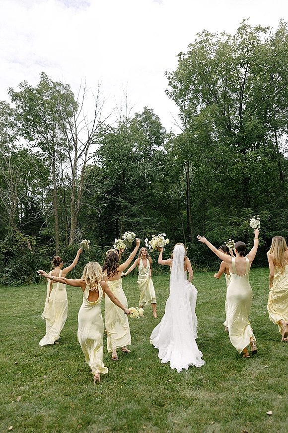 Bridesmaids photo of the bride with bridesmaids in satin yellow dresses holding bouquets on a grassy lawn by trees at the forest edge