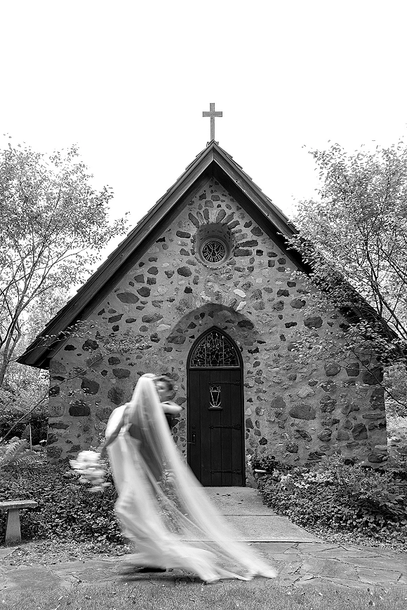 Bridal portrait in a black and white wedding photo of a bride walking with long veil and bouquet past a stone chapel door and cross