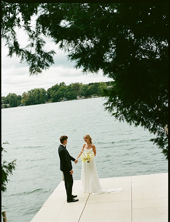 Couple portrait at a lake wedding portrait on a dock, bride in strapless dress and veil holding a white bouquet beside groom in black tux