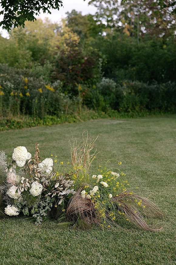 Ceremony aisle florals in a grounded cluster of white hydrangeas, roses, yellow wildflowers and dried grasses on a garden lawn
