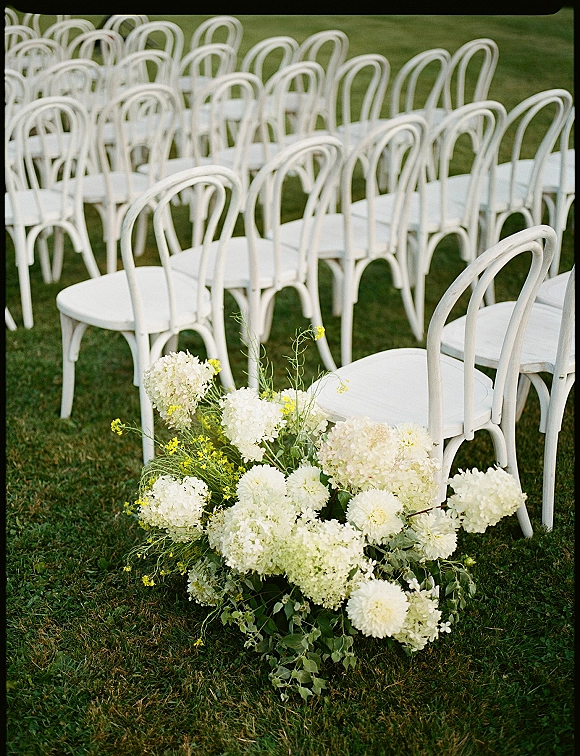 Ceremony seating with outdoor ceremony chairs in neat rows of white bentwood chairs beside white and green aisle florals on a grass lawn