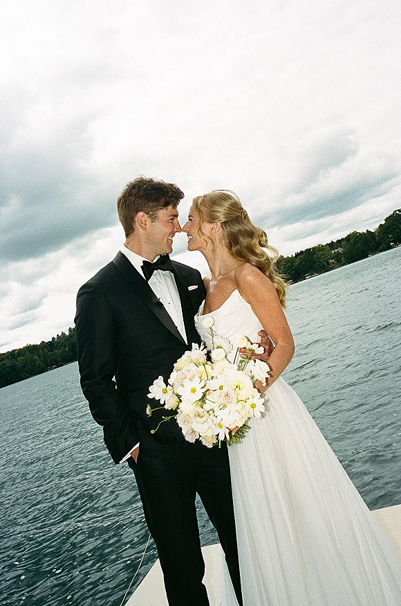 Couple portrait of bride and groom standing nose to nose on a dock, bride holding white bouquet beside a cloudy lake backdrop