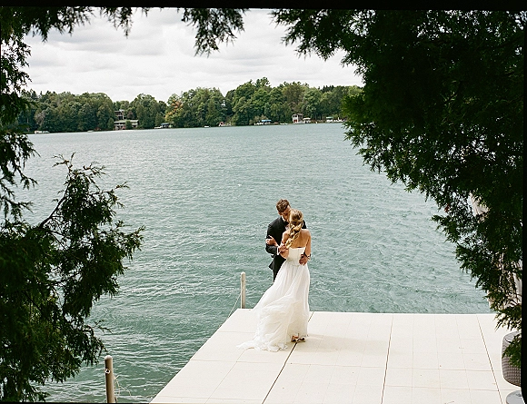 Wedding couple portrait of bride and groom kissing on a dock, her veil trailing behind as a cloudy lake and evergreens frame them