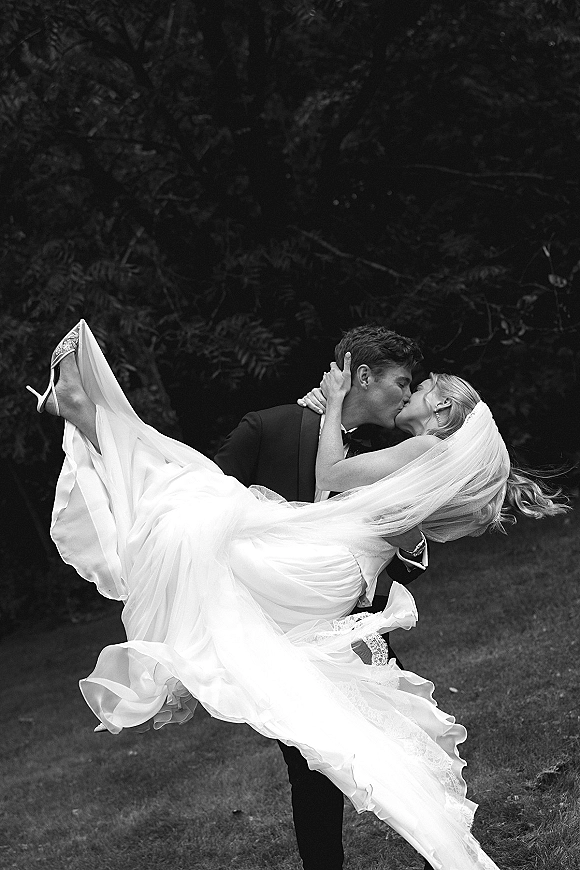 Wedding kiss portrait of groom lifting bride in a dipped kiss, her veil flowing behind strapless dress on a garden lawn with trees
