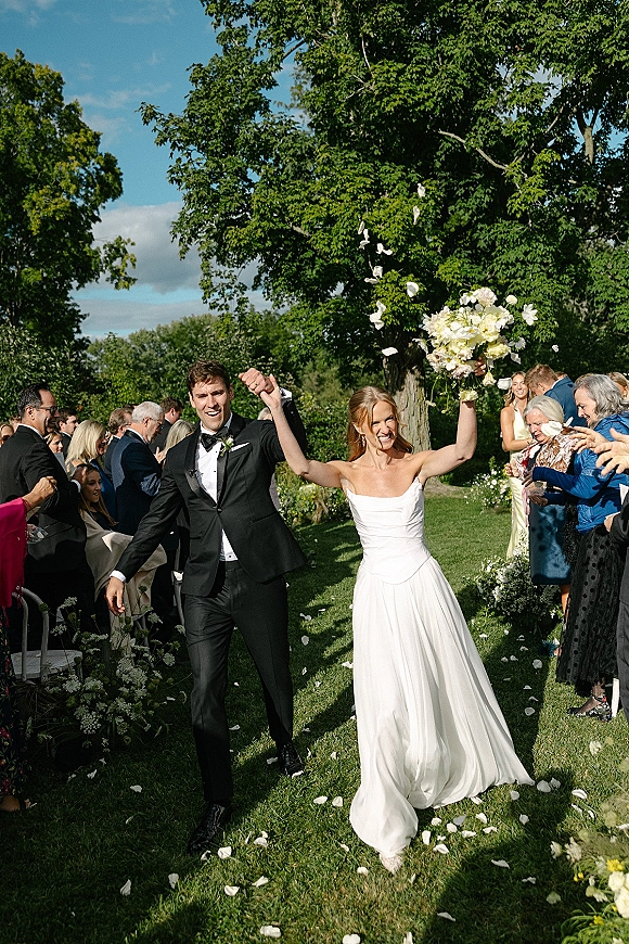 Wedding recessional as bride and groom walk the aisle holding hands, bride lifting bouquet amid rose petal toss on sunny lawn with guests cheering