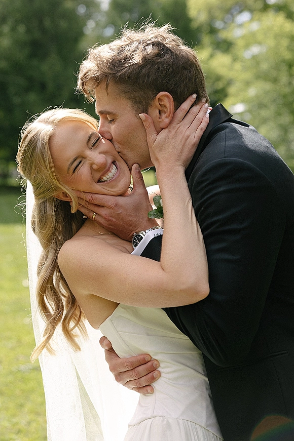 Wedding kiss portrait of bride and groom kissing as she laughs, holding his face, veil flowing in sunlit greenery outdoors