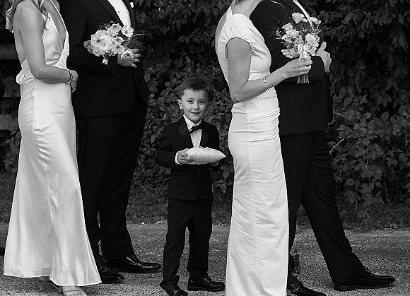 Ring bearer portrait of a little boy in a tuxedo holding a ring pillow, smiling beside bridesmaids with bouquets on a garden path