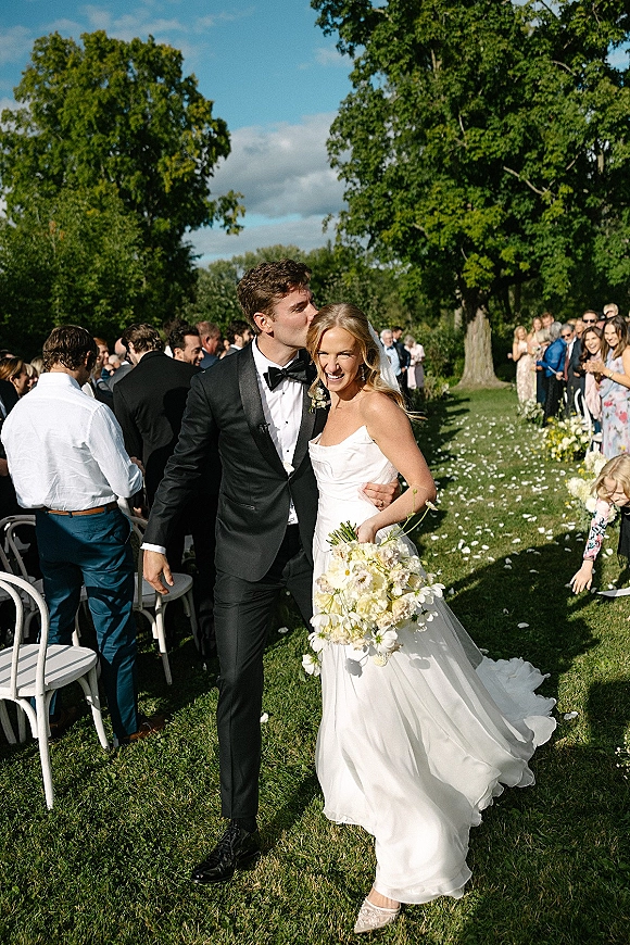 Recessional moment as bride and groom walk the aisle at an outdoor wedding recessional, groom kissing her forehead with petals and guests on lawn