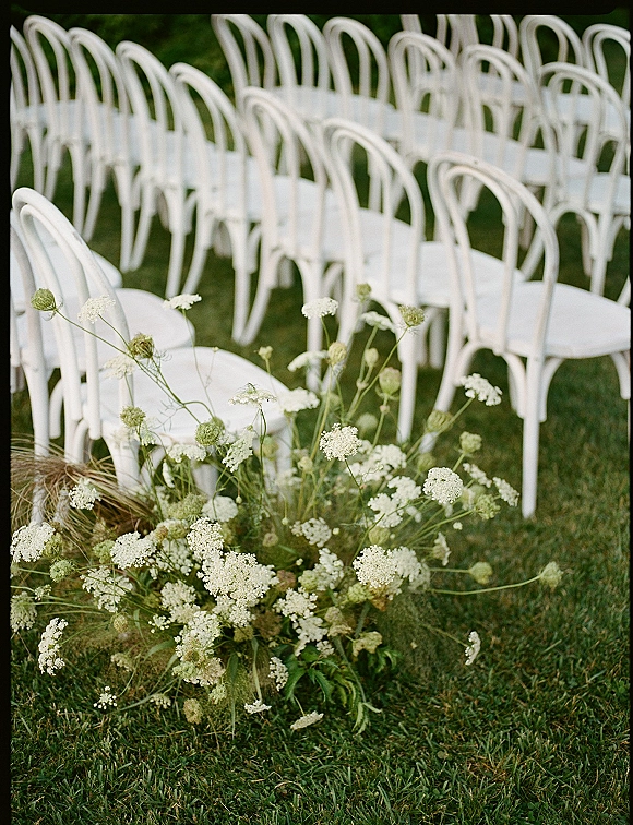 Ceremony seating with outdoor ceremony chairs in neat rows of white chairs, framed by low floral aisle arrangement on a garden lawn