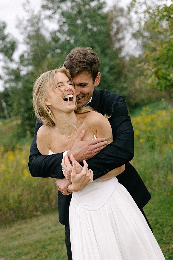 Couple portrait of bride and groom laughing as he hugs her from behind, showing her strapless wedding dress and jewelry in a meadow setting