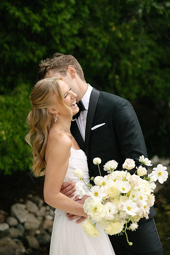 Couple portrait of bride and groom embrace as he kisses her cheek, bride holding white bouquet against green foliage and stone wall backdrop