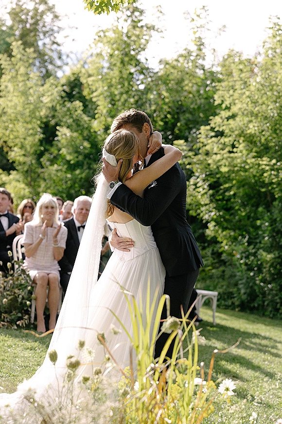 Wedding kiss portrait of bride and groom kissing, her long veil flowing as seated guests watch in a lush garden ceremony setting