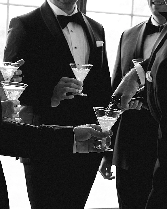 Wedding cocktail toast as groomsmen in tuxedos raise martini glasses with a champagne bottle by a large window in soft indoor light